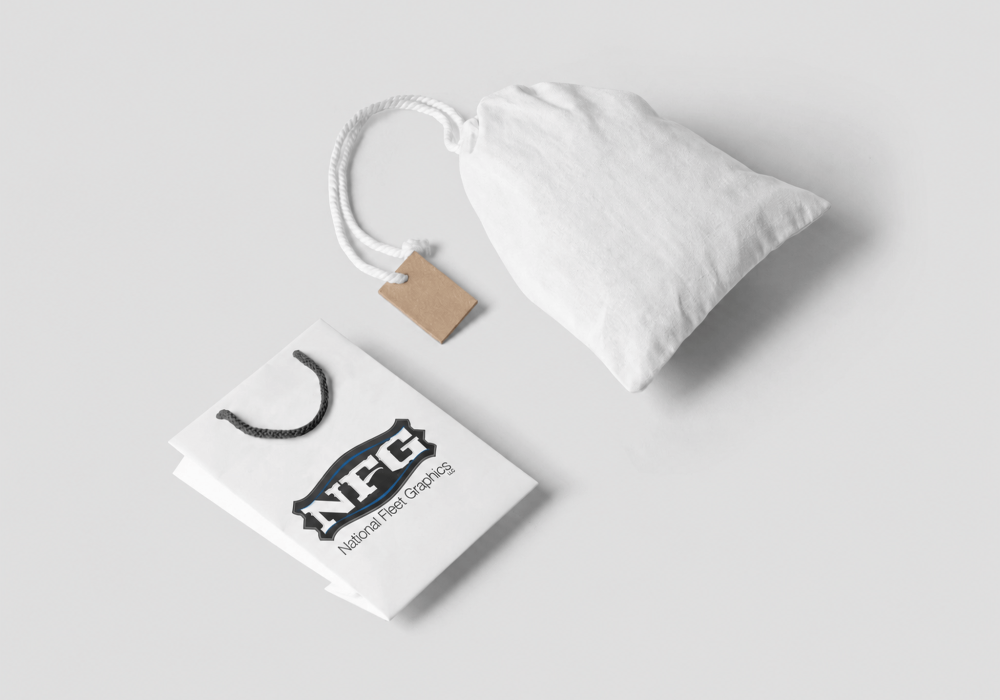 Flat lay of a white drawstring cloth pouch with a brown tag, next to a white shopping bag with black rope handles and a blue shield logo on it.